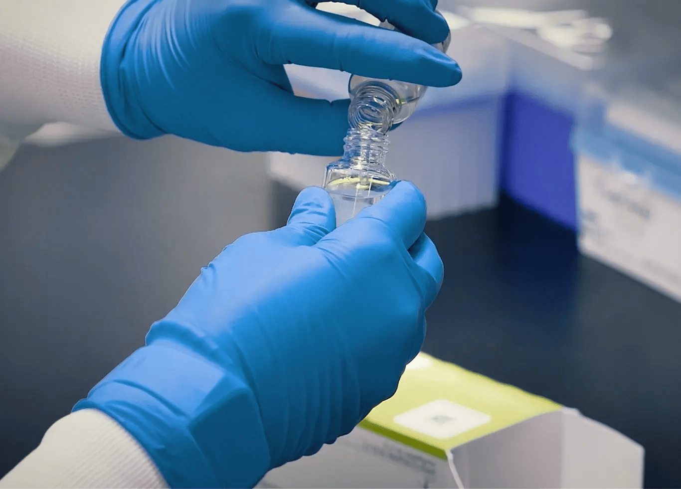 Photo of a person's hands wearing blue laboratory gloves and pouring a liquid reagent into another bottle. The branded green top of the HemosIL Reagent box can be seen in the background.